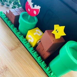 May include: A decorative tray featuring elements from the Super Mario video game series. Includes a red and white Piranha Plant, green pipes, brown brick blocks, a yellow question mark block, and a yellow star. The tray is green with a black border.