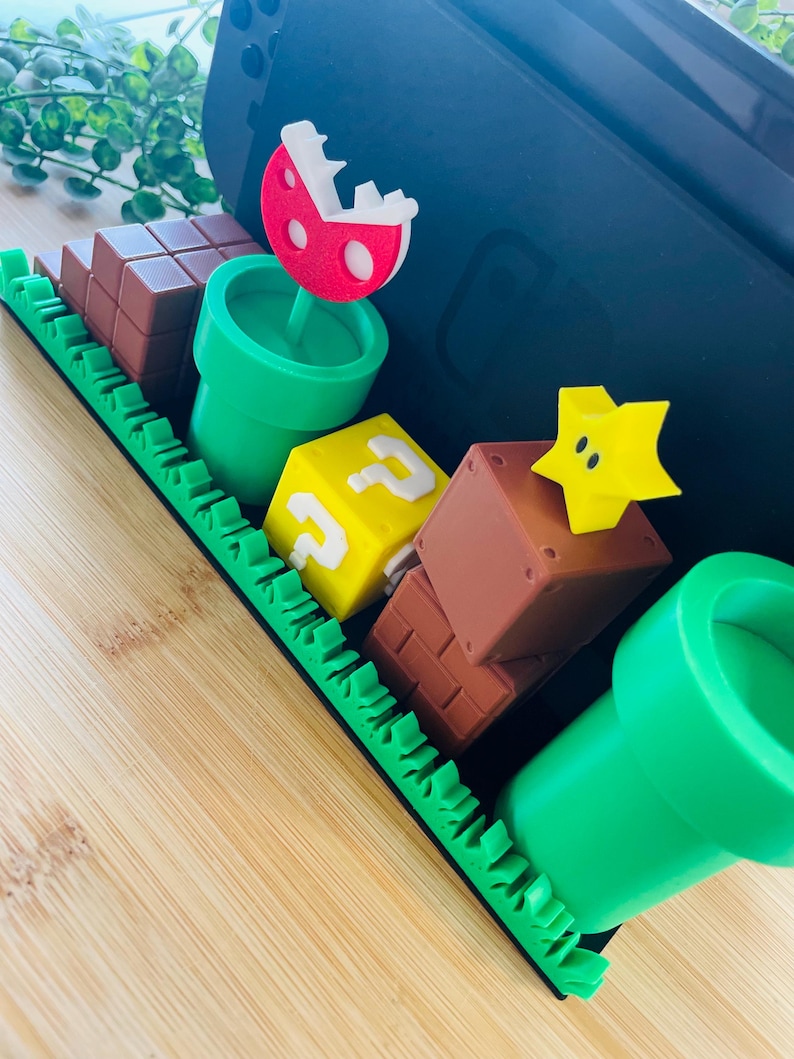 May include: A decorative tray featuring elements from the Super Mario video game series. Includes a red and white Piranha Plant, green pipes, brown brick blocks, a yellow question mark block, and a yellow star. The tray is green with a black border.