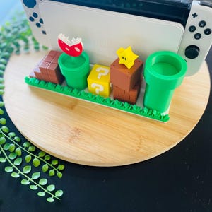 May include: A white Nintendo Switch console rests on a decorative stand featuring iconic elements from the Super Mario video game series. The stand includes green pipes, question mark blocks, and a red and white Piranha Plant.