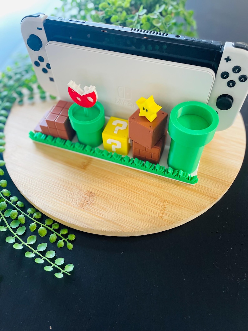 May include: A white Nintendo Switch console rests on a decorative stand featuring iconic elements from the Super Mario video game series. The stand includes green pipes, question mark blocks, and a red and white Piranha Plant.