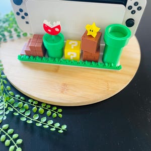 May include: A white and black Nintendo Switch console is displayed on a wooden stand with a Super Mario Bros. theme. The stand features green pipes, question mark blocks, brown brick blocks, a star, and a Piranha Plant.