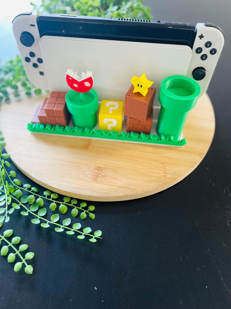 May include: A white and black Nintendo Switch console is displayed on a wooden stand with a Super Mario Bros. theme. The stand features green pipes, question mark blocks, brown brick blocks, a star, and a Piranha Plant.