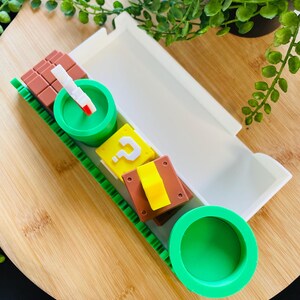 May include: A white tray with a green and brown decorative border, containing various colorful, themed objects. Includes a green pipe, a brown block, a yellow question mark block, and a white and red object. The tray sits on a wooden surface.