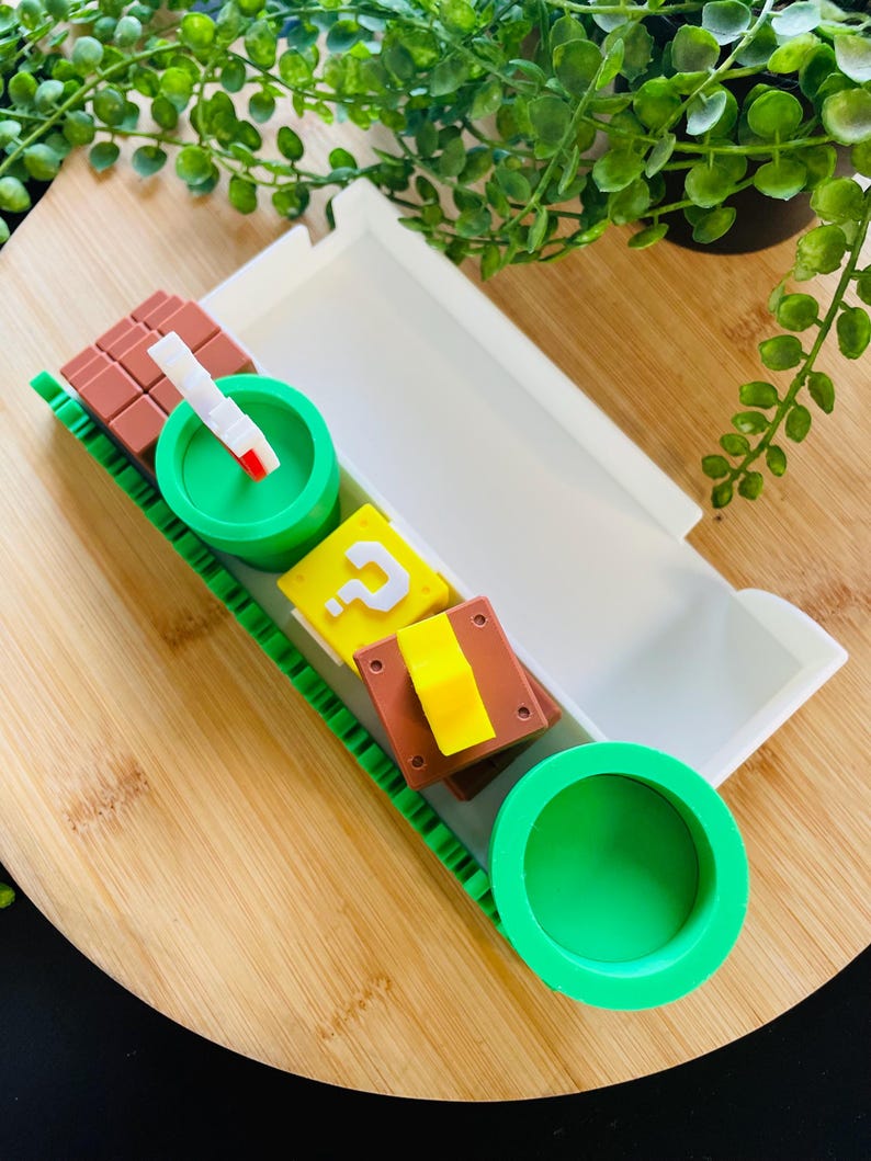 May include: A white tray with a green and brown decorative border, containing various colorful, themed objects. Includes a green pipe, a brown block, a yellow question mark block, and a white and red object. The tray sits on a wooden surface.