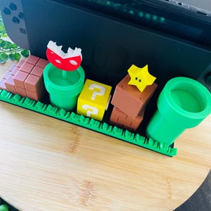 May include: A black Nintendo Switch console is the backdrop for a colorful diorama of Super Mario Bros. game elements. The scene includes brown brick blocks, green pipes, a yellow question mark block, a red and white Piranha Plant, and a yellow star.
