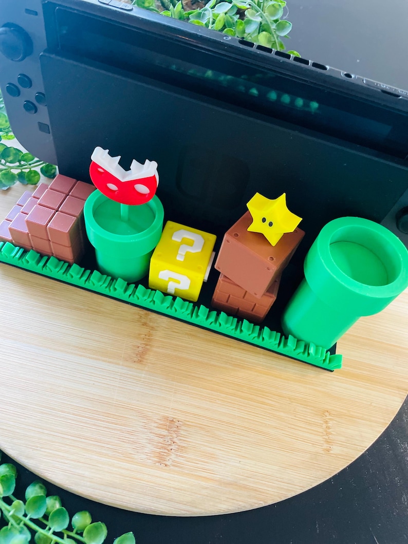 May include: A black Nintendo Switch console is the backdrop for a colorful diorama of Super Mario Bros. game elements. The scene includes brown brick blocks, green pipes, a yellow question mark block, a red and white Piranha Plant, and a yellow star.