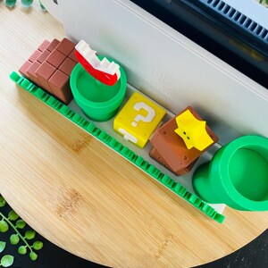 May include: A white game console with a black screen is next to several colorful, miniature Super Mario-themed figures. The figures include a red and white boat, a yellow question mark block, and a brown block.