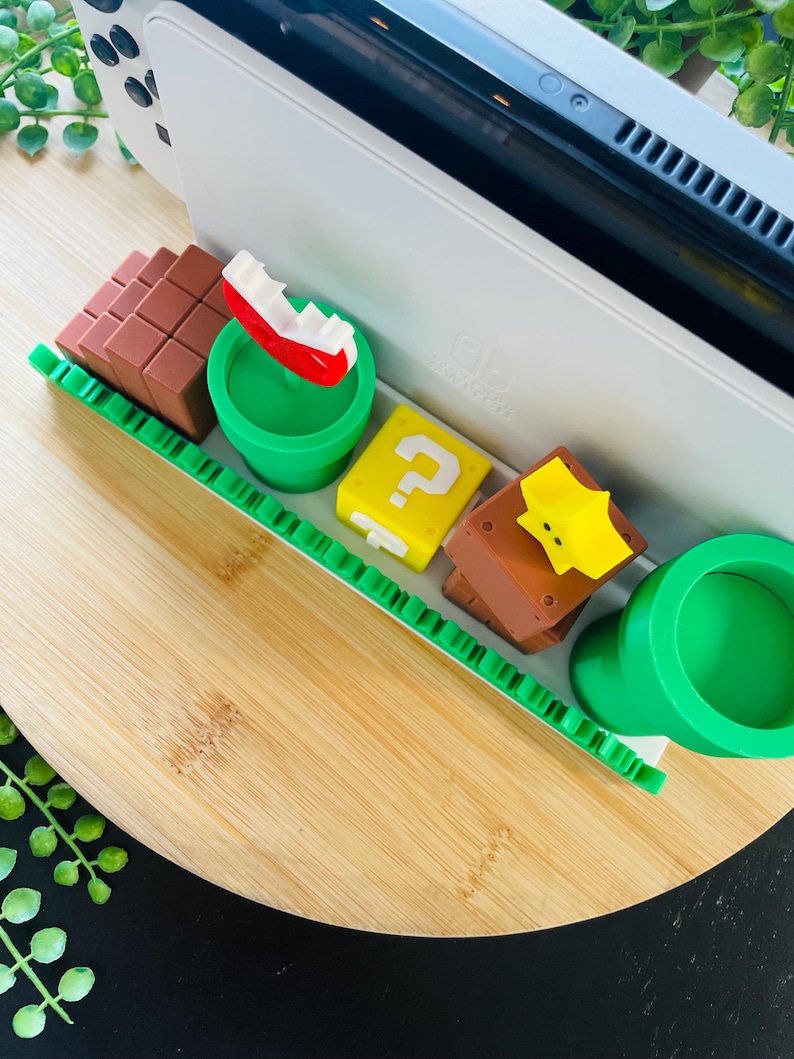 May include: A white game console with a black screen is next to several colorful, miniature Super Mario-themed figures. The figures include a red and white boat, a yellow question mark block, and a brown block.