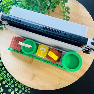 May include: A black and white Nintendo Switch console rests on a white charging dock, with a green and brown themed game cartridge holder. The holder features a green pipe, question mark block, and other game-themed elements.