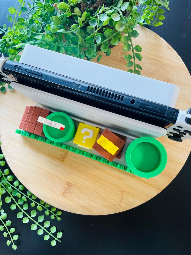 May include: A black and white Nintendo Switch console rests on a white charging dock, with a green and brown themed game cartridge holder. The holder features a green pipe, question mark block, and other game-themed elements.