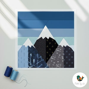 May include: A framed fabric art piece depicting a mountain range. The artwork features a white frame and a patchwork design with shades of blue, grey, and white. The mountains have white peaks and the sky is a gradient of blues. Two spools of thread are in the lower left.