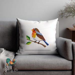 May include: White square throw pillow featuring a geometric design of a bird perched on a branch. The bird has a brown, orange, and blue color scheme. The pillow is on a gray couch.
