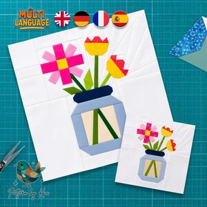 Könnte beinhalten: Ein Quiltblock-Muster mit einer hellblauen Vase mit dunkelblauem Rand, die eine rosa Blume und zwei gelb-rote Tulpen mit grünen Stielen auf weißem Hintergrund enthält. Eine kleinere Version desselben Blumendesigns ist ebenfalls zu sehen.