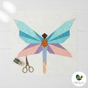 May include: A colourful dragonfly quilt block with blue, purple, and peach wings. The dragonfly is centred on a white background with geometric patterns. A spool of thread and small scissors are next to the quilt block.