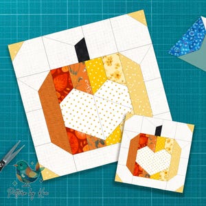May include: Two pumpkin-themed quilt blocks. The larger block features a pumpkin design with orange, yellow, and white fabrics. The smaller block mirrors the design. The blocks are on a teal cutting mat. The text "Pattern by Kia" is visible.