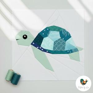 Op de afbeelding: Een geometrisch quiltblok met een zeeschildpadontwerp. De schildpadschild is samengesteld uit verschillende blauwe en groene stoffen. De kop en vinnen zijn lichtgroen. Twee klosjes garen staan in de linker benedenhoek.