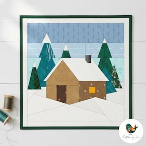 May include: A quilted wall hanging featuring a winter cabin scene. The cabin is brown with a snow-covered roof, set against a backdrop of blue and green trees and a snowy landscape. The piece is framed in a dark green border.
