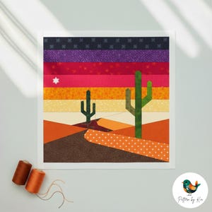 Peut inclure: Une œuvre d'art en tissu représentant un coucher de soleil dans le désert avec des cactus. Le ciel est un dégradé de rayures violettes, rouges, oranges et jaunes. Deux cactus verts se dressent au premier plan. L'œuvre est encadrée en blanc, avec deux bobines de fil en bas à gauche.