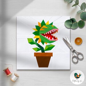 May include: A paper piecing quilt block pattern featuring a green and yellow carnivorous plant in a brown pot. The plant has a wide open mouth with red interior and sharp white teeth. The design is set against a white background with visible seam lines.