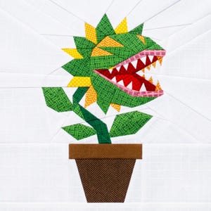 May include: A fabric quilt block pattern featuring a carnivorous plant with green, yellow, and red patterned fabric pieces. The plant has sharp white teeth, an open mouth, and green leaves, all set in a brown pot against a white grid background.