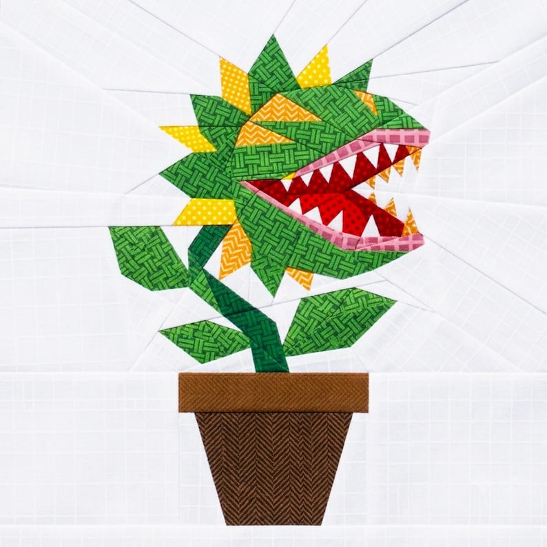 May include: A fabric quilt block pattern featuring a carnivorous plant with green, yellow, and red patterned fabric pieces. The plant has sharp white teeth, an open mouth, and green leaves, all set in a brown pot against a white grid background.