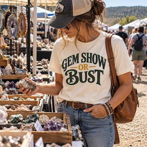 May include: A cream-colored short-sleeve t-shirt featuring the dark green and gold text GEM SHOW OR BUST printed on the front. The shirt is worn with blue denim jeans, a brown leather belt, a brown leather backpack, and a baseball cap.