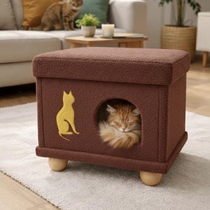 Puede incluir: Una casa para gatos marrón con una entrada redonda y una silueta de gato amarilla. Un gato jengibre está durmiendo dentro. La casa para gatos está sobre una alfombra blanca, con un gato en un sofá en el fondo.