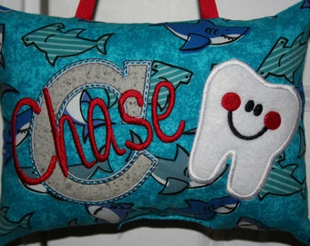Può includere: Un cuscino blu con un dente bianco e il nome "Chase" ricamato in rosso. Il cuscino ha un motivo a squalo.