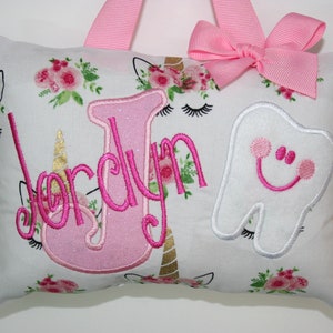 May include: A white pillow with a pink floral pattern and a pink unicorn and tooth design. The pillow has a pink bow and the name "Jordyn" embroidered on it.