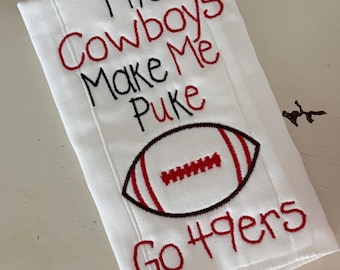 Può includere: Un canovaccio bianco con testo ricamato che recita The Cowboys Make Me Puke e Go 49ers, accompagnato da un grafico ricamato di un pallone da football con dettagli in filo rosso e nero.