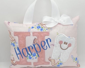 Può includere: Un cuscino rosa con un manico e un fiocco bianchi. Il cuscino ha una grande lettera rosa "H" con il nome "Harper" ricamato in blu. Un dente bianco con un viso sorridente si trova sul lato destro del cuscino.