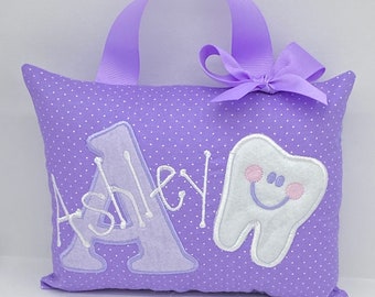 Può includere: Cuscino viola con pois bianchi, un dente bianco con un viso sorridente rosa e il nome "Ashley" ricamato in bianco. Il cuscino ha un manico viola.