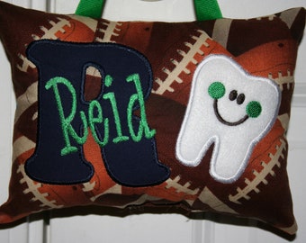 Può includere: Un cuscino marrone e beige con un motivo a pallone da calcio. Il cuscino ha una grande lettera blu "R" con il nome "Reid" ricamato in verde. C'è un dente bianco con un viso sorridente e occhi verdi sul cuscino.