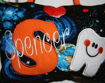 Può includere: Un cuscino nero e blu a tema spaziale. Il cuscino ha una grande S arancione con il nome "Spencer" ricamato in bianco. C'è un dente bianco con un viso sorridente e guance rosse sul lato destro del cuscino.