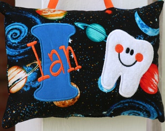 Può includere: Un cuscino nero e blu a tema spaziale. Il cuscino ha una grande lettera "I" blu con il nome "Ian" ricamato in arancione. Un dente bianco con un viso sorridente è anche ricamato sul cuscino.