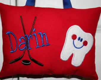 Può includere: Cuscino rosso con bordo blu e un dente bianco con un viso sorridente, due bastoni da hockey incrociati, un disco nero e il nome "Darin" ricamato in blu.