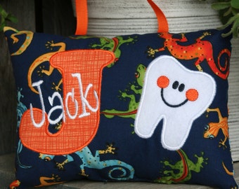 Può includere: Un cuscino blu con un dente bianco e una lettera J arancione. Il cuscino ha un motivo a lucertola verde e arancione. La lettera J è ricamata con il nome "Jack".