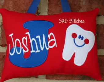 Può includere: Cuscino rosso con ricamo blu e bianco. Il cuscino presenta una grande lettera J blu e il nome Joshua. Un dente bianco con un viso sorridente è ricamato sul lato destro del cuscino. Il testo "S&D Stitches" è ricamato nell'angolo in alto a destra.
