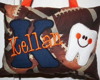 Può includere: Un cuscino marrone e beige con un motivo a pallone da calcio. Il cuscino ha una grande lettera K blu con il nome "Kellan" ricamato in arancione. Un dente bianco con un viso sorridente e guance arancioni si trova sul lato destro del cuscino.
