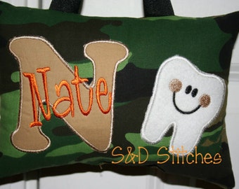 Può includere: Un cuscino di mimetica verde con una lettera N ricamata in marrone e il nome "Nate" in arancione. Un dente bianco con un viso sorridente e guance marroni è ricamato sul lato destro del cuscino. Il testo "S&D Stitches" è ricamato in arancione nella parte inferiore del cuscino.
