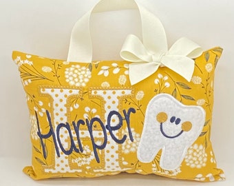 Può includere: Un cuscino giallo con un motivo floreale bianco e un dente bianco con un viso sorridente ricamato. Il cuscino ha anche il nome "Harper" ricamato in filo blu navy. Il cuscino ha un anello di nastro bianco per appenderlo.