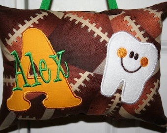 Può includere: Un cuscino per fatina dei denti a tema football marrone e beige con il nome "Alex" ricamato in verde su una lettera "A" gialla. Un'applicazione di dente sorridente è sul lato destro. Un nastro verde è attaccato per appenderlo.