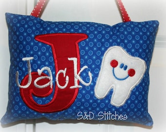 Può includere: Un cuscino blu e rosso con pois bianchi. Il cuscino ha il nome "Jack" ricamato in rosso con un contorno bianco. Un dente bianco con un viso sorridente rosso è ricamato sul lato destro del cuscino. Il cuscino ha un anello di nastro rosso e bianco per appenderlo. Il testo "S&D Stitches" è ricamato nell'angolo in basso a destra.