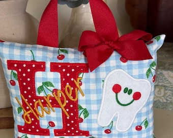 Può includere: Un piccolo cuscino rettangolare con motivo a quadretti vichy azzurro e bianco e stampa di ciliegie rosse. Presenta un manico e un fiocco in nastro rosso, una grande lettera H rossa a pois, il nome Harper in giallo e un'applicazione a dente.