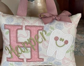 Può includere: Un cuscino rettangolare in tessuto con manico e fiocco in nastro rosa polvere. Il cuscino presenta una stampa floreale, una grande lettera H rosa ricamata, il nome Harper in corsivo verde e un'applicazione di un dente bianco sorridente.