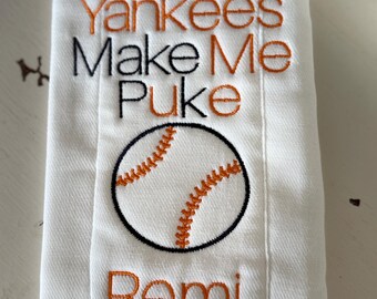 Può includere: Asciugamano bianco a tema baseball. Il testo ricamato recita: "The Yankees Make Me Puke" in nero e arancione. Una grafica di baseball è sotto il testo e il nome "Remi" è ricamato in arancione.