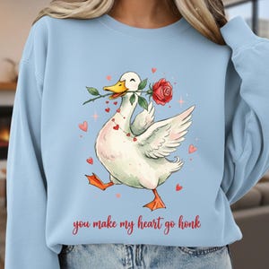 May include: Pale blue sweatshirt with a cartoon duck carrying a red rose. The duck has a heart necklace and is surrounded by hearts. The text "you make my heart go honk" is printed below the duck in a handwritten style font.