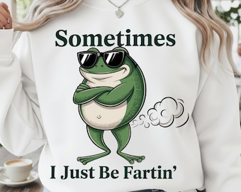 May include: White sweatshirt with the text "Sometimes I Just Be Fartin'" and a cartoon frog wearing sunglasses. The frog is green with a white belly and arms crossed. A cloud of gas is behind the frog.