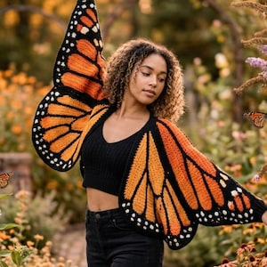 Könnte beinhalten: Ein schwarzes Häkeltop und schwarze Jeans werden mit einer Häkel-Schmetterlingsflügel-Strickjacke getragen. Die Strickjacke hat orangefarbene und schwarze Flügel mit weißen Punkten an den Rändern, die an einen Monarchfalter erinnern.