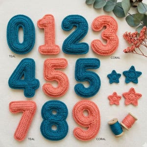 May include: A collection of handmade crochet numbers and stars in teal and coral colors. The numbers 0 through 9 are displayed, along with two teal and two coral stars. Teal and coral thread spools are also visible.